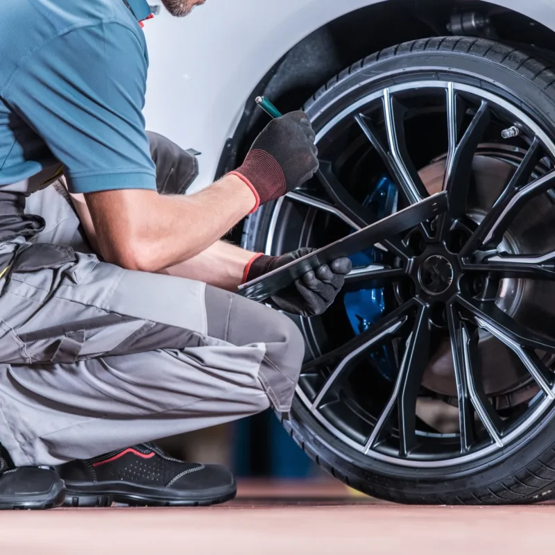 A mechanic in gray overalls and work gloves kneels beside a white car's front wheel, holding a pen and clipboard while inspecting the tire and blue brake caliper.