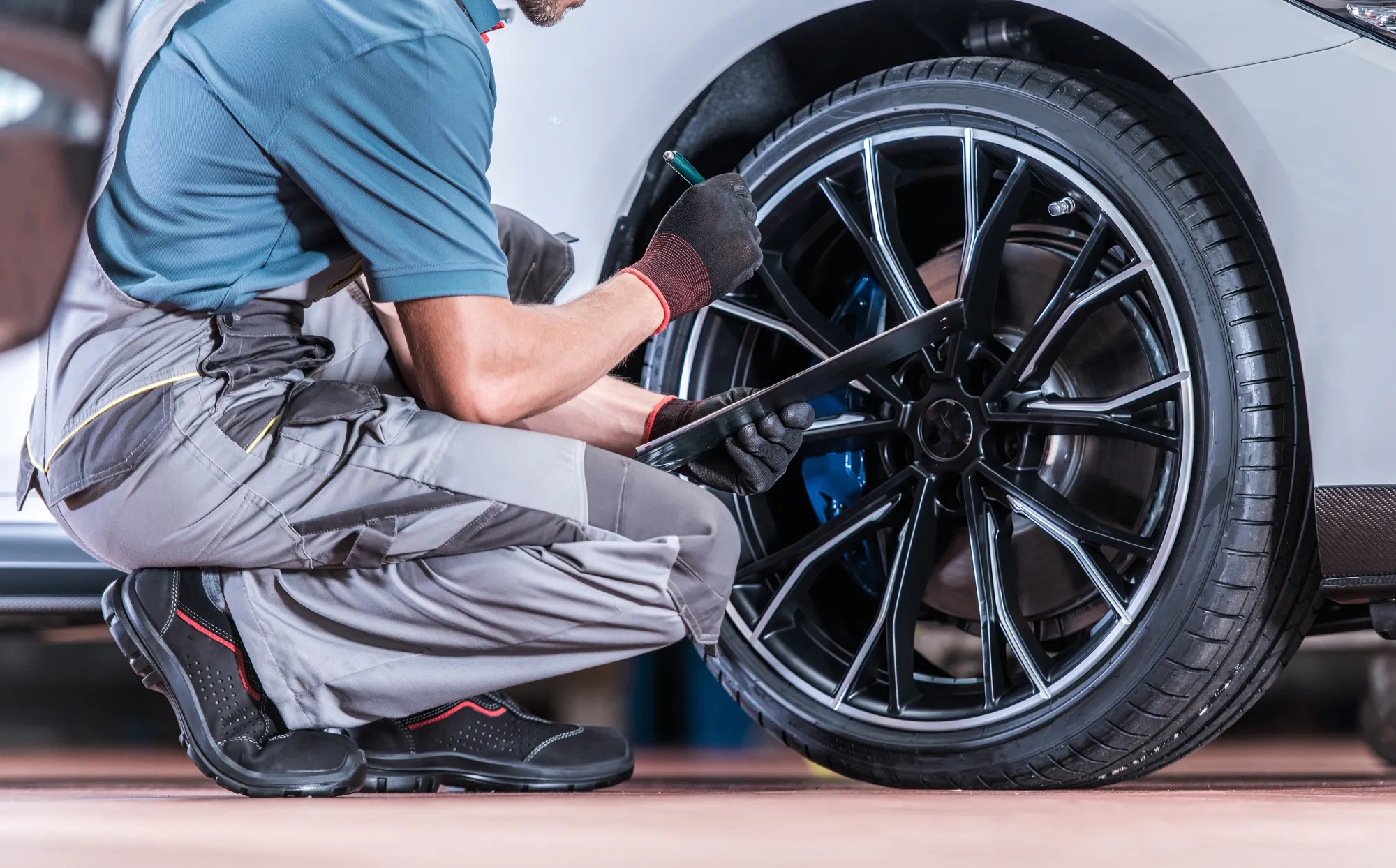 A mechanic in gray overalls and work gloves kneels beside a white car's front wheel, holding a pen and clipboard while inspecting the tire and blue brake caliper.