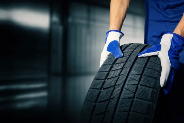 A mechanic in blue coveralls and work gloves rolls a new black tire across a garage floor.