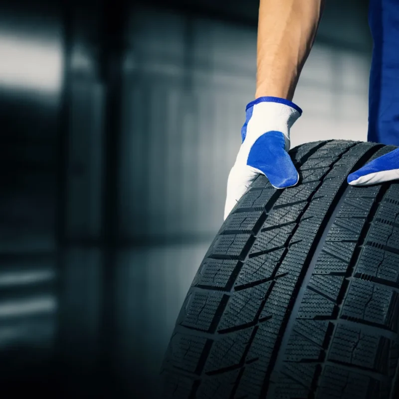 A mechanic in blue coveralls and work gloves rolls a new black tire across a garage floor.