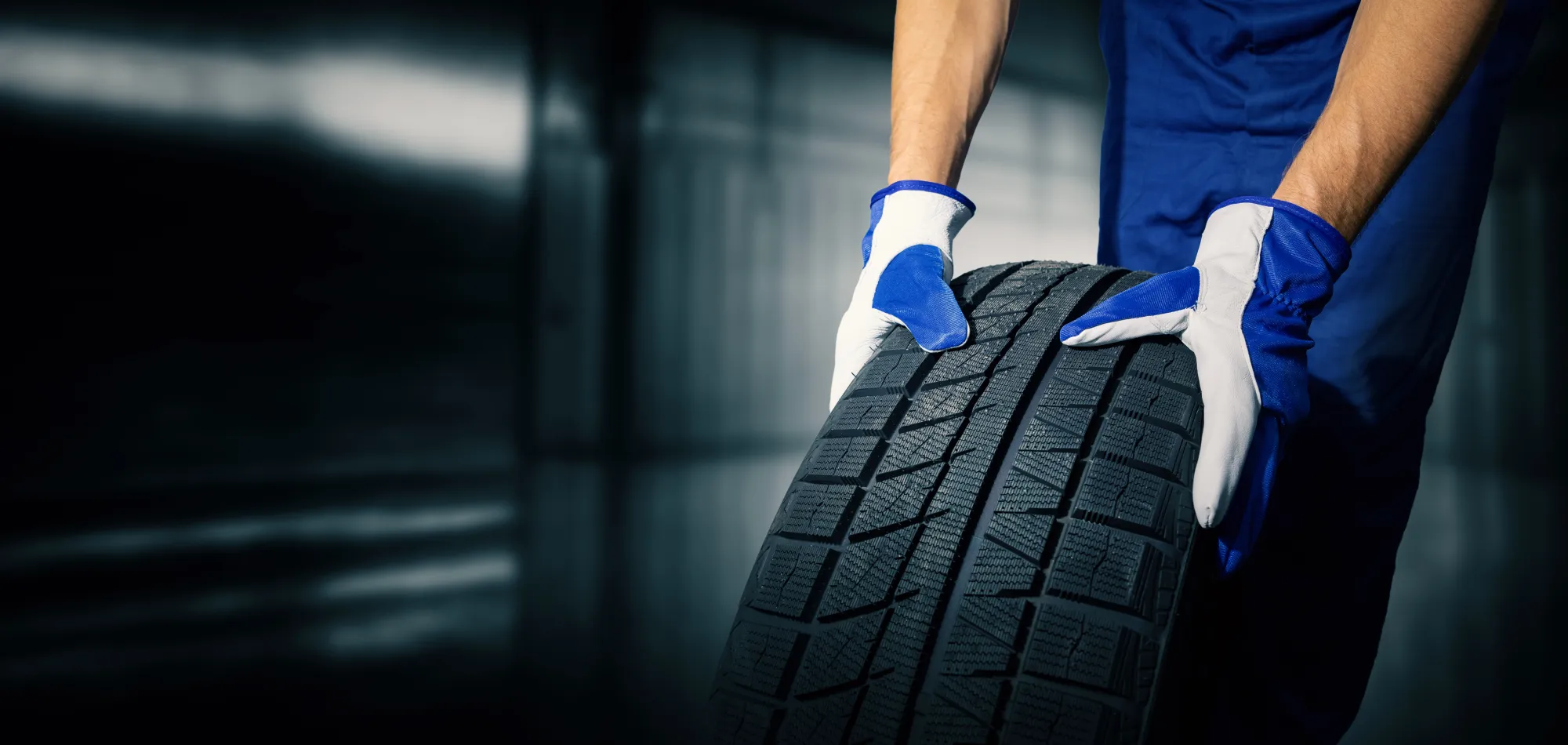A mechanic in blue coveralls and work gloves rolls a new black tire across a garage floor.
