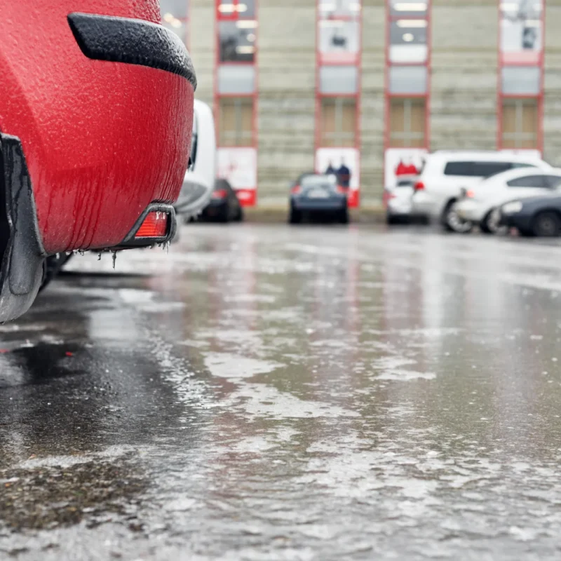 Icicles hang from the rear bumper of a red car parked on an icy, wet lot in winter.
