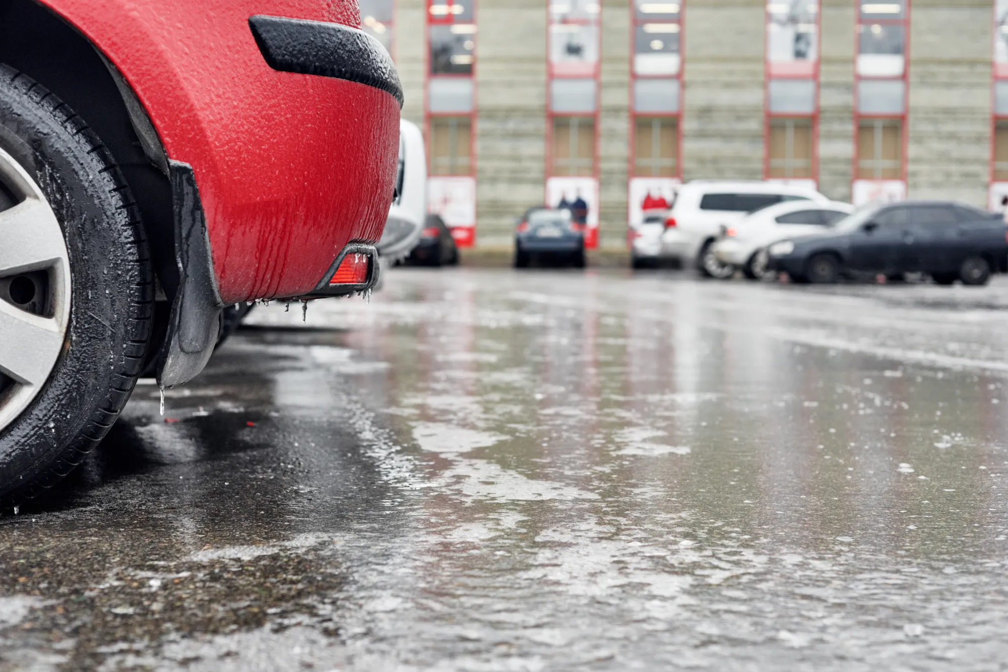 Icicles hang from the rear bumper of a red car parked on an icy, wet lot in winter.