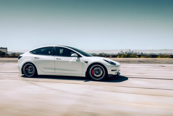 A white Tesla Model 3 sedan with dark alloy wheels and red brake calipers drives at speed across open pavement with a city skyline in the distance.