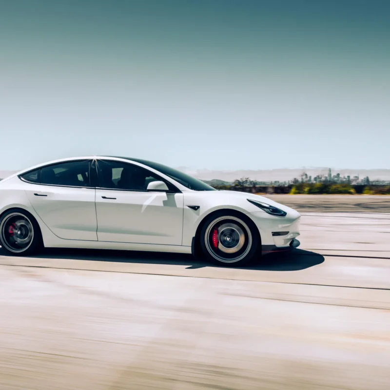 A white Tesla Model 3 sedan with dark alloy wheels and red brake calipers drives at speed across open pavement with a city skyline in the distance.