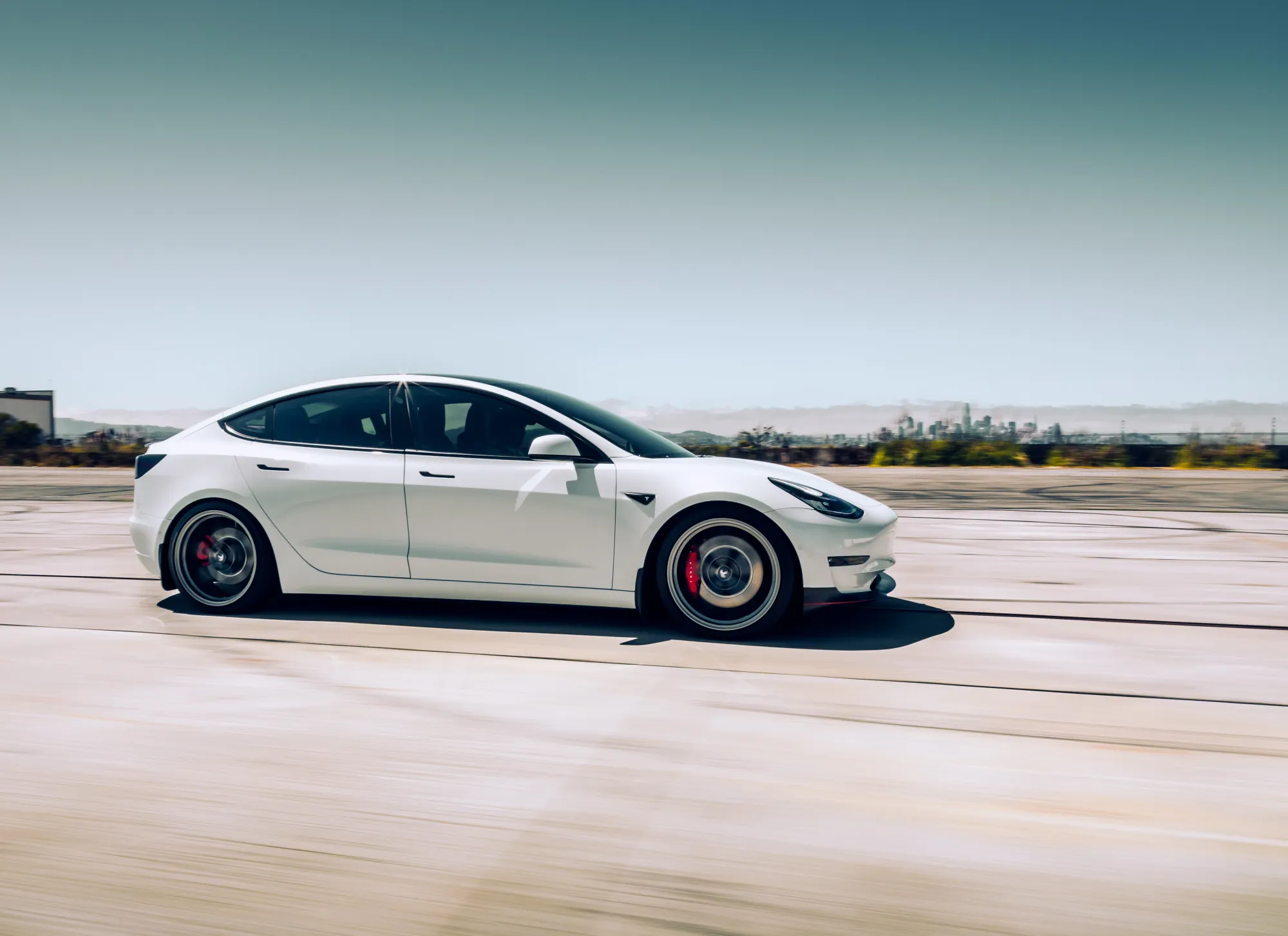 A white Tesla Model 3 sedan with dark alloy wheels and red brake calipers drives at speed across open pavement with a city skyline in the distance.