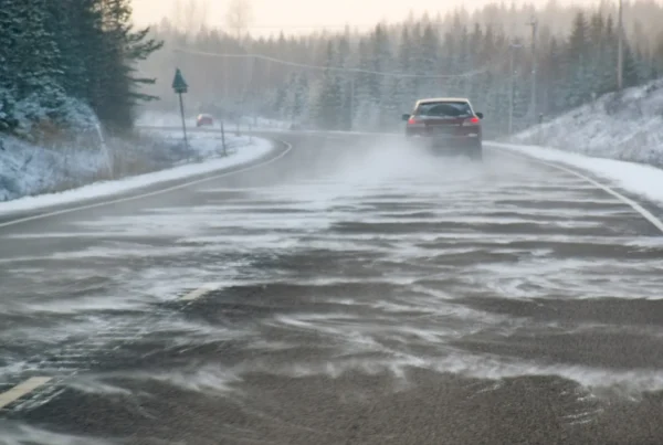 A red car driving on a snowy highway as wind blows snow across the icy road between forested shoulders.