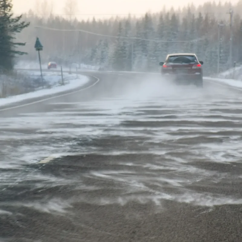 A red car driving on a snowy highway as wind blows snow across the icy road between forested shoulders.
