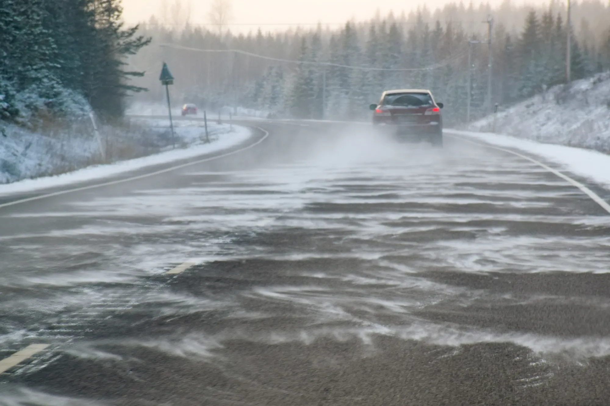 A red car driving on a snowy highway as wind blows snow across the icy road between forested shoulders.