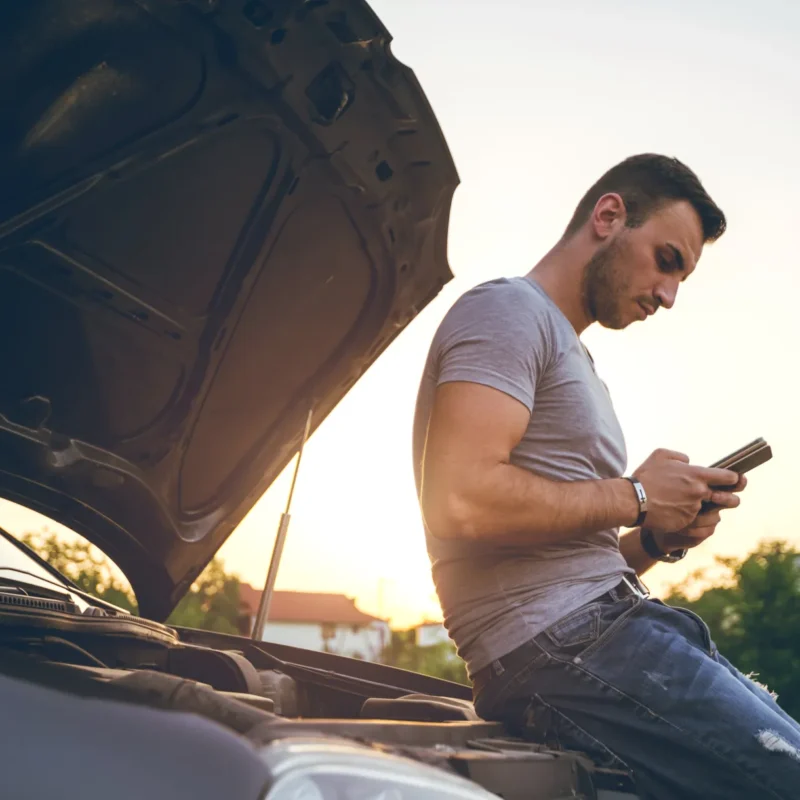 A man leans on the front of a car with the hood open, checking his phone at sunset beside the road.
