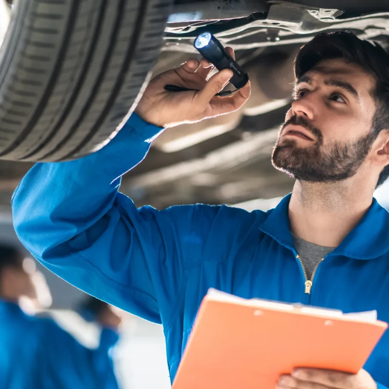 A mechanic in blue coveralls holding a clipboard shines a flashlight at the underside of a car raised on a lift in a repair shop.