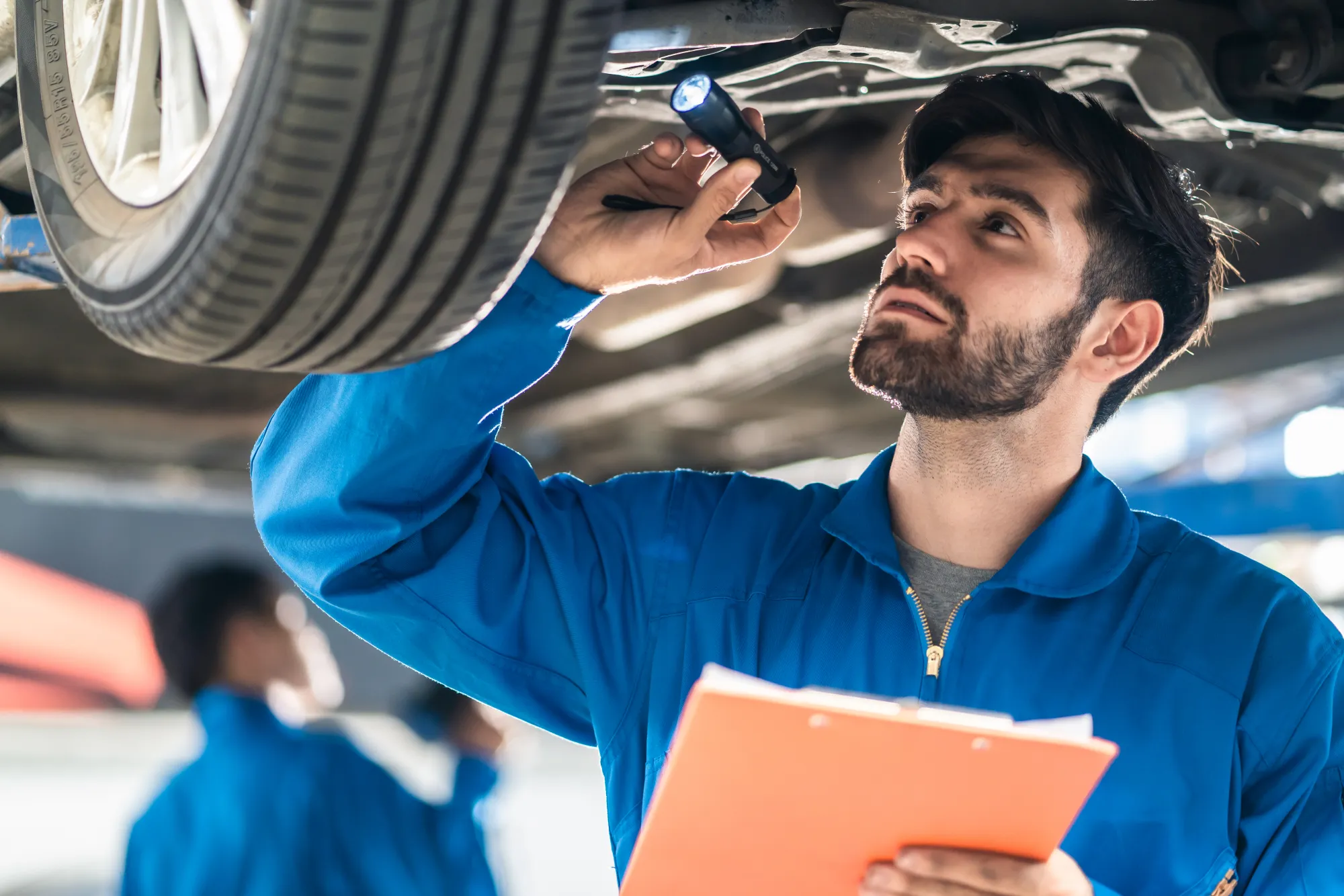 A mechanic in blue coveralls holding a clipboard shines a flashlight at the underside of a car raised on a lift in a repair shop.