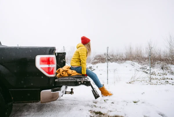 A woman in a yellow winter jacket and red knit hat sits on the open tailgate of a black pickup truck parked in a snowy field.