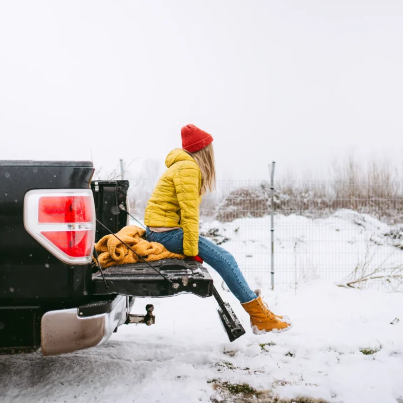 A woman in a yellow winter jacket and red knit hat sits on the open tailgate of a black pickup truck parked in a snowy field.