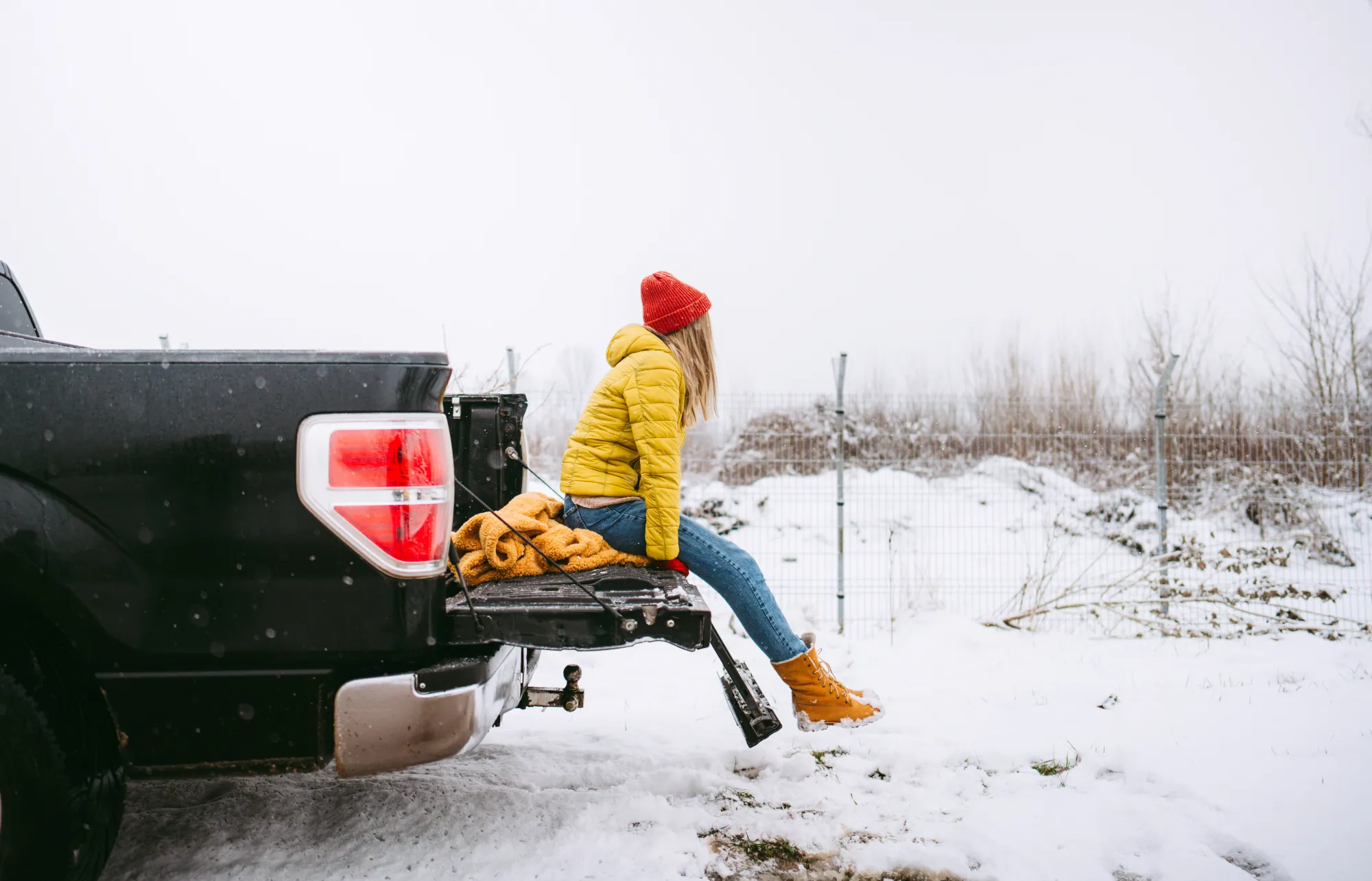 A woman in a yellow winter jacket and red knit hat sits on the open tailgate of a black pickup truck parked in a snowy field.
